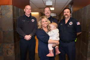 Blonde woman holding infant standing in front of three paramedics