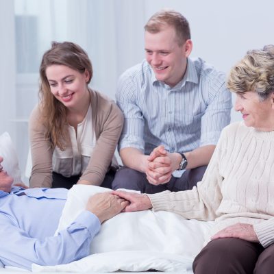 Old man lying in a white hospital bed, surrounded by his smiling relatives