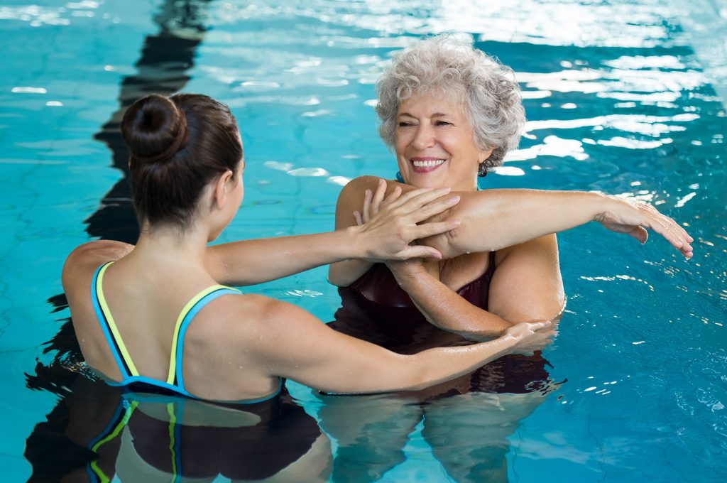Female water therapy instructor assisting an elderly female student in stretching while in the pool