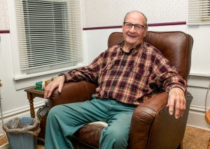 Elderly gentleman in plaid shirt sits in brown leather armchair
