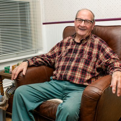 Elderly gentleman in plaid shirt sits in brown leather armchair